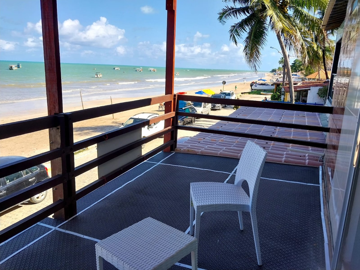 A spacious balcony overlooks the beach, featuring two white chairs positioned beside a railing. The sandy shore is visible, with calm waves and fishing boats in the distance. Lush palm trees provide shade, complementing the tranquil coastal scenery.