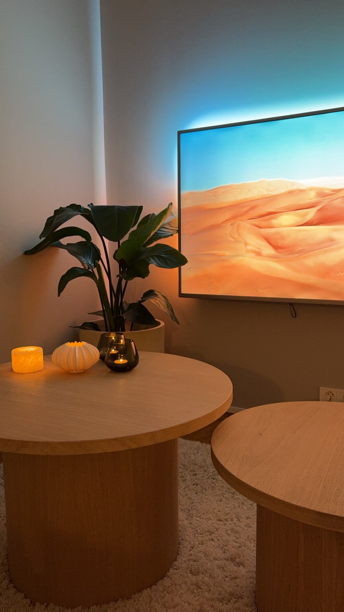A cozy living area features two light wooden tables positioned near a wall-mounted TV displaying a desert landscape. A vibrant green plant sits beside the tables, while soft lighting is provided by a candle and a decorative pumpkin on the table.