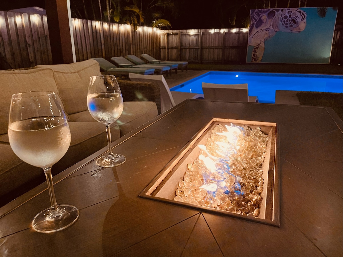 A close-up view of a table featuring two glasses filled with clear liquid, positioned next to a fire pit with sparkling glass stones. In the background, a well-lit pool is visible, reflecting the blue water, with lounge chairs arranged nearby and soft lighting enhancing the evening atmosphere.
