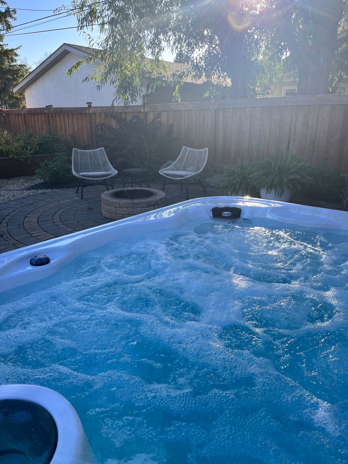 A hot tub is positioned nearby, surrounded by a well-maintained patio and lush greenery. Two modern chairs are situated across from a circular fire pit, all under soft sunlight filtering through the trees.