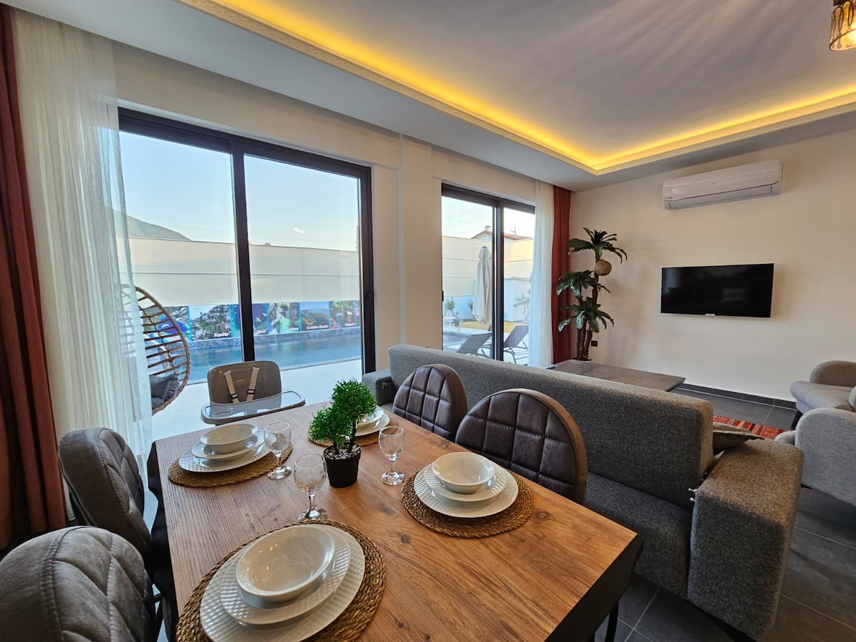 A dining area is set with neatly arranged dinnerware atop a wooden table. Large windows allow natural light to fill the space, showcasing views of the terrace. Comfortable seating is available nearby, complemented by greenery adding a touch of nature to the interior.