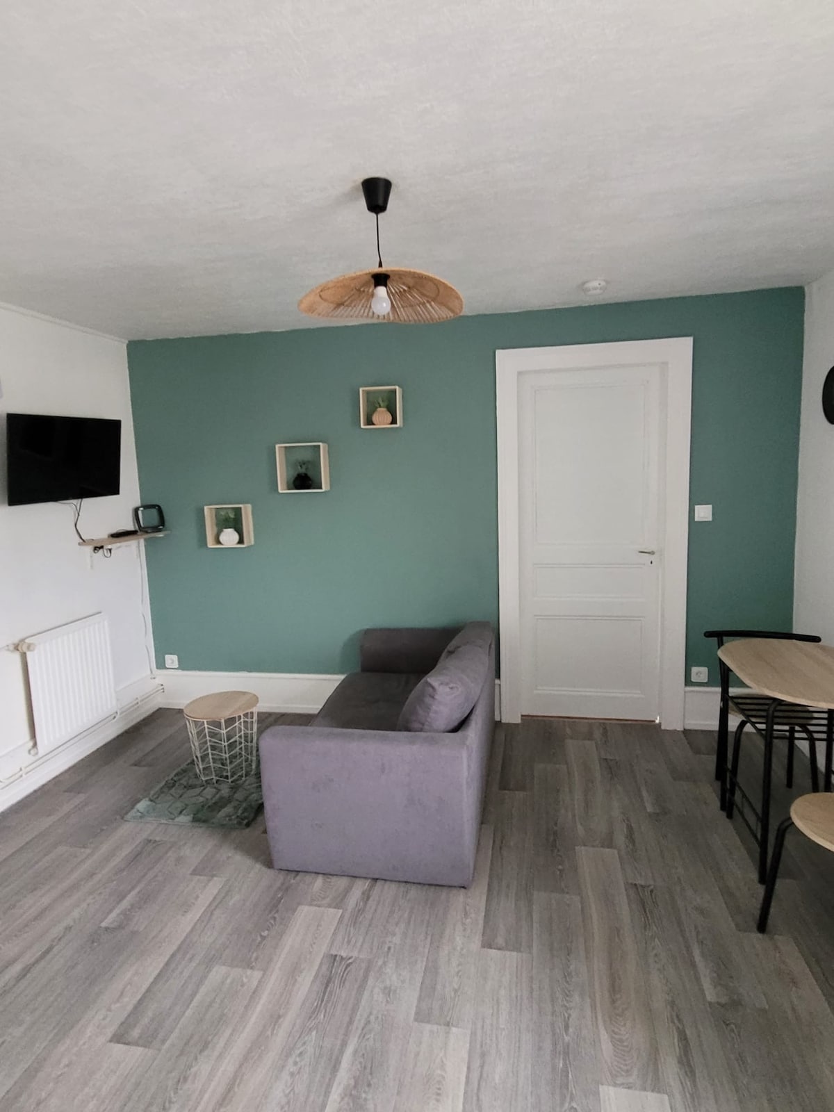 A living area is presented with a light green accent wall. A grey sofa sits beside a small round table and two black bar stools. A mounted television is positioned on the wall, with decorative shelves displaying various items, creating a simple yet functional space.