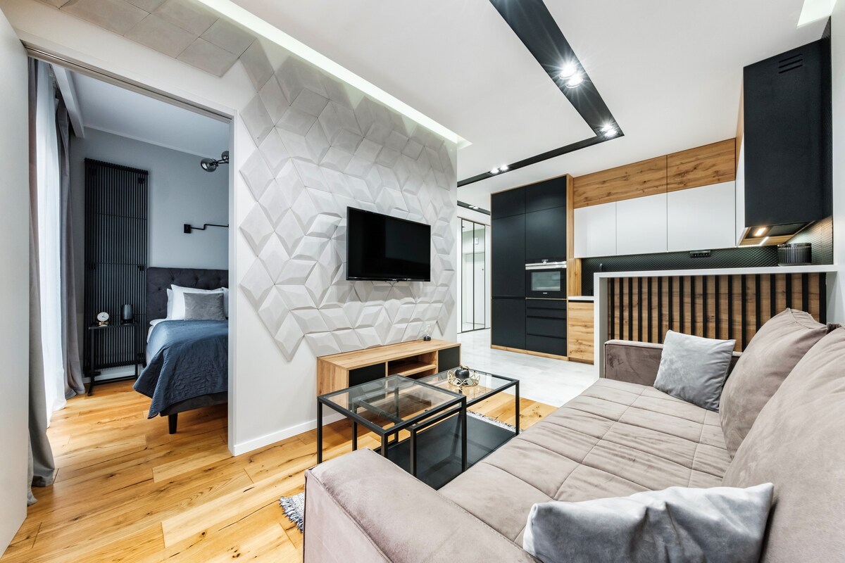 A modern living area is highlighted by a textured accent wall and a sleek television mounted at eye level. A sectional sofa faces a glass coffee table, while an open doorway leads to a bedroom featuring a comfortable bed with dark bedding.