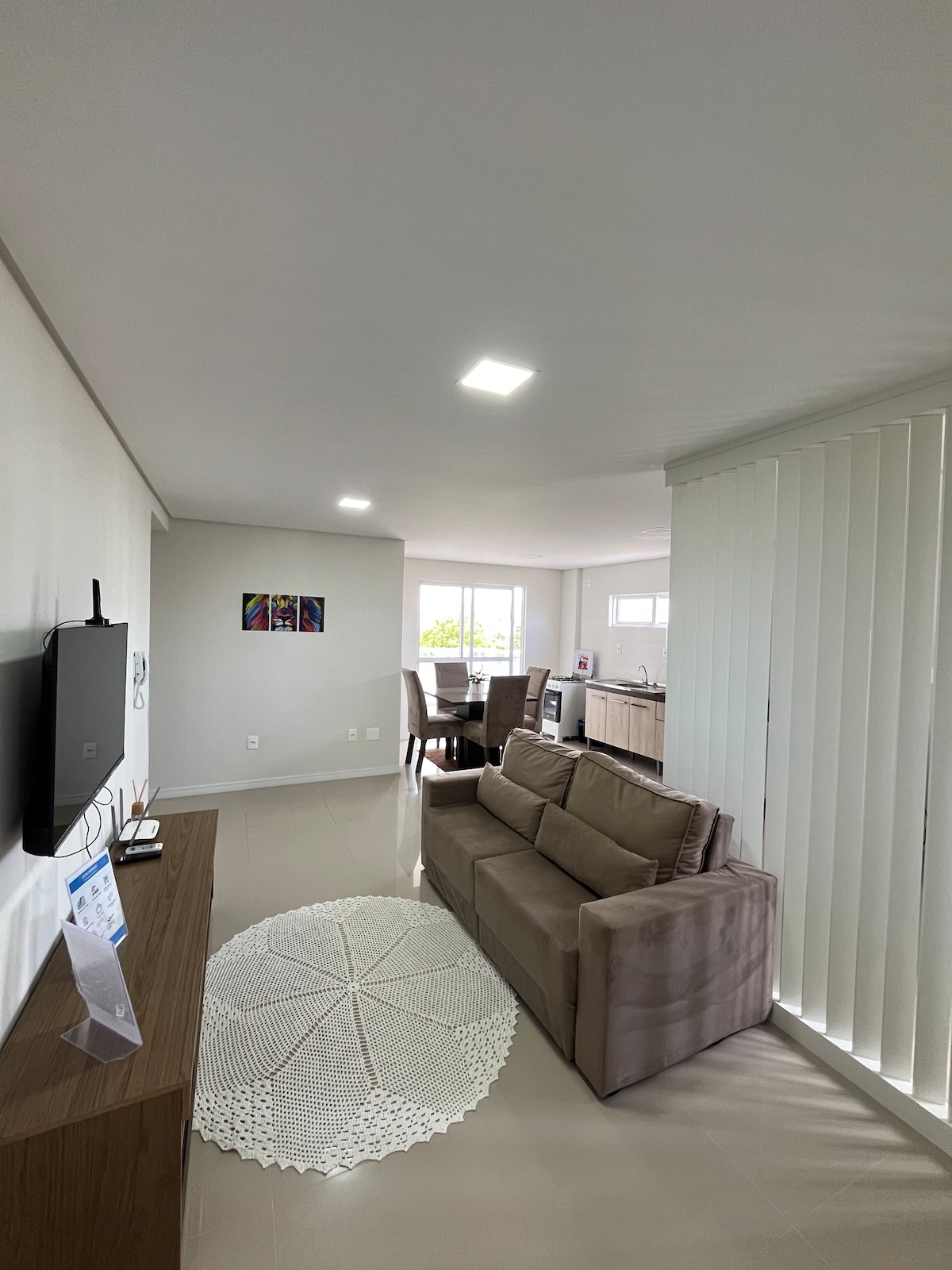 A well-lit living area features a comfortable three-seater sofa facing a wall-mounted television. Natural light enters through a large window, complemented by vertical blinds. A dining area with a glass table and four chairs is visible in the background, alongside a modern kitchen.