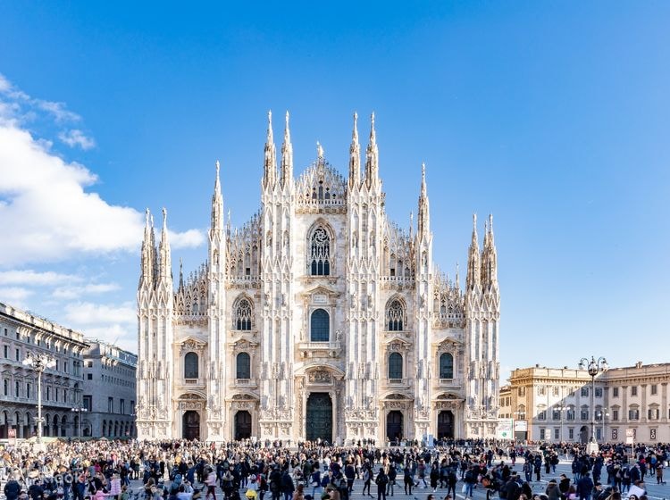 A grand cathedral with intricate Gothic architecture dominates the scene, featuring tall spires and ornate details. Crowds of visitors gather in the plaza, set against a clear blue sky. Nearby buildings contribute to the urban atmosphere surrounding the cathedral.
