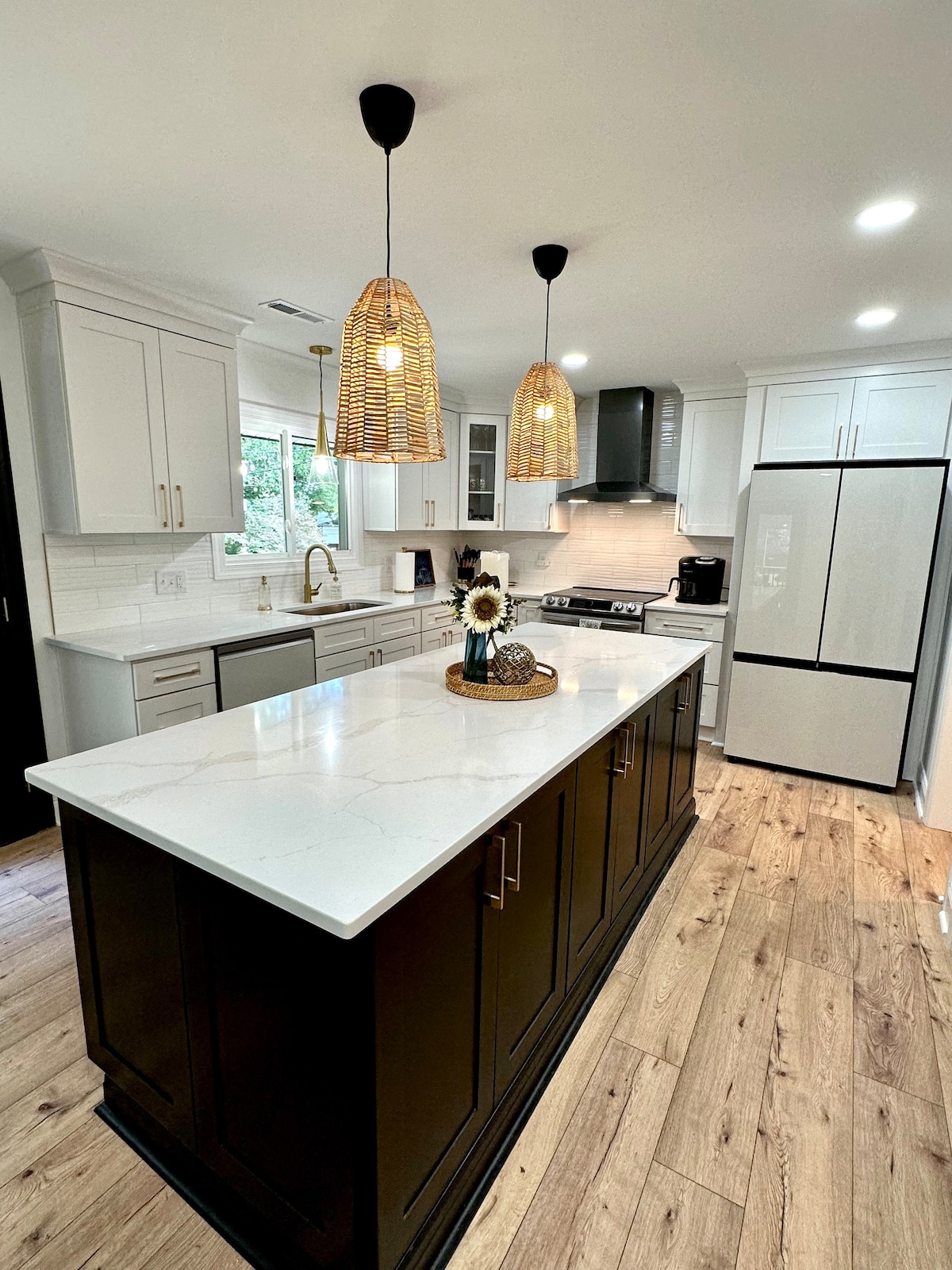 A modern kitchen exhibits sleek cabinetry in light and dark tones. A spacious island features a white countertop with a decorative arrangement of sunflowers. Pendant lights hang above, while stainless steel appliances and ample natural light contribute to the functional design.
