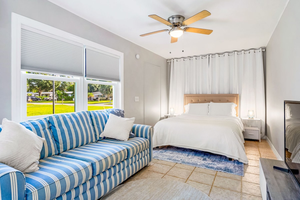 A bright studio space features a spacious bed with neutral bedding, complemented by a soft, striped couch. Natural light enters through two windows, and light-colored curtains hang softly against a light gray wall. A ceiling fan adds a functional touch to the inviting room.