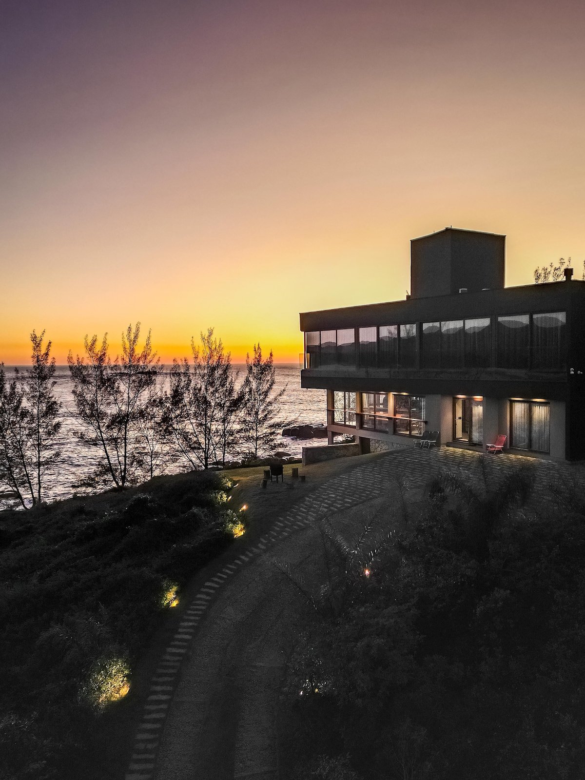 A modern house is positioned near the ocean, with large glass windows reflecting the warm colors of a sunset. Lush greenery surrounds the property, and soft lighting highlights the pathway leading to the entrance.