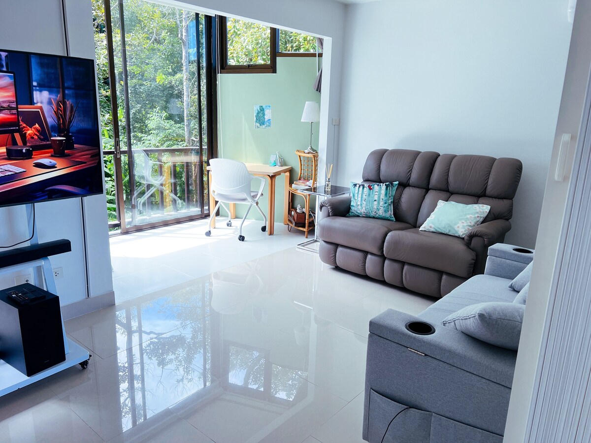 The living room features a comfortable recliner sofa and a light grey couch facing a large digital TV. A spacious window allows natural light to fill the room, with views of greenery visible outside. A small desk and chair are positioned near the window.