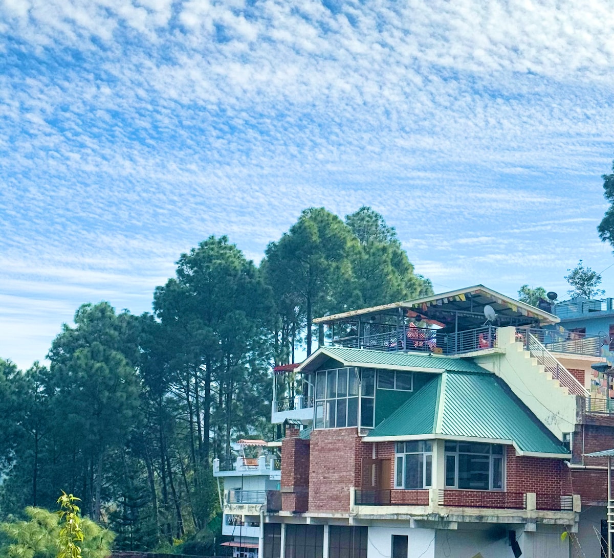 A multi-level villa is surrounded by lush pine trees, showcasing its distinct architecture. The upper floor features a spacious terrace with seating and panoramic views of the sky, while large windows reflect the natural light throughout the structure.