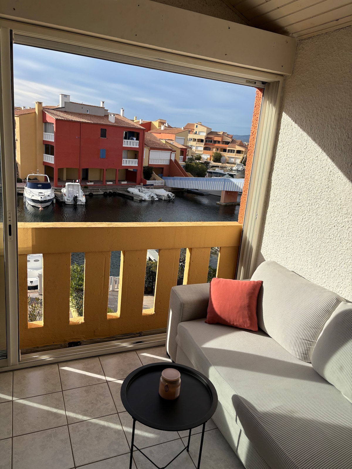 A bright corner of the studio features a light-colored sofa and a small black side table. Large sliding glass doors open to a balcony overlooking a waterway lined with boats and colorful buildings, allowing natural light to enhance the space.