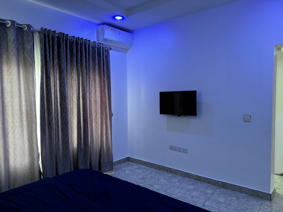 A cozy bedroom is presented, featuring soft blue lighting. A wall-mounted television is positioned across from a spacious bed. Flowing curtains softly cover a window, allowing ambient light to filter in, while a sleek air conditioning unit is installed above for comfort.