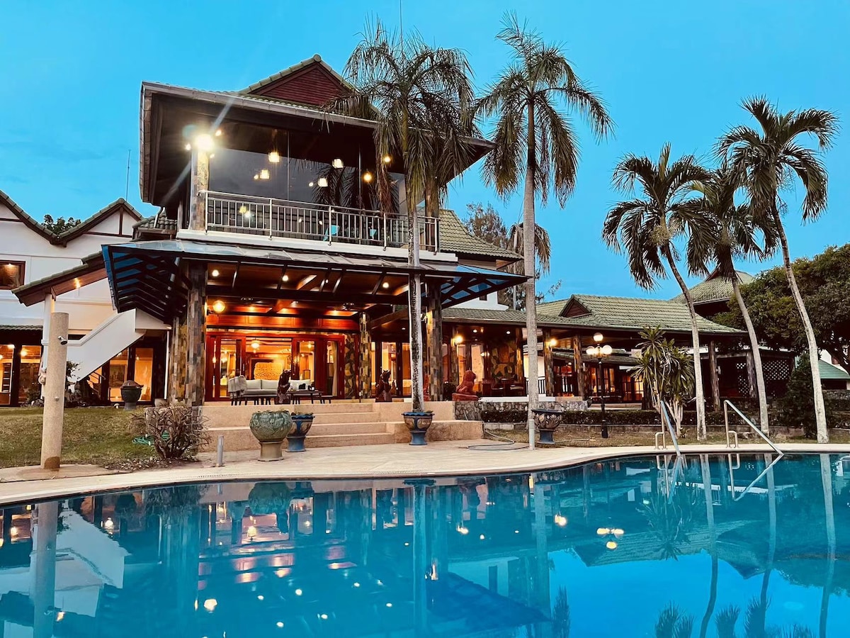 The image features a luxurious two-story villa surrounded by palm trees, reflecting in a clear blue swimming pool. The outdoor area is illuminated, highlighting a spacious terrace with seating arrangements and vibrant landscaping, enhancing the connection between indoor and outdoor spaces.
