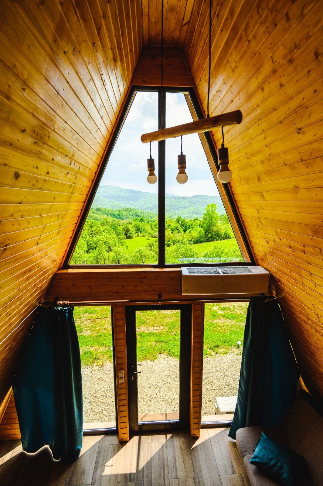 A striking A-frame interior is featured, showcasing wooden walls and a tall triangular window that provides a panoramic view of the lush greenery outside. Soft curtains frame the window, and a glimpse of outdoor space is visible through the glass doors.