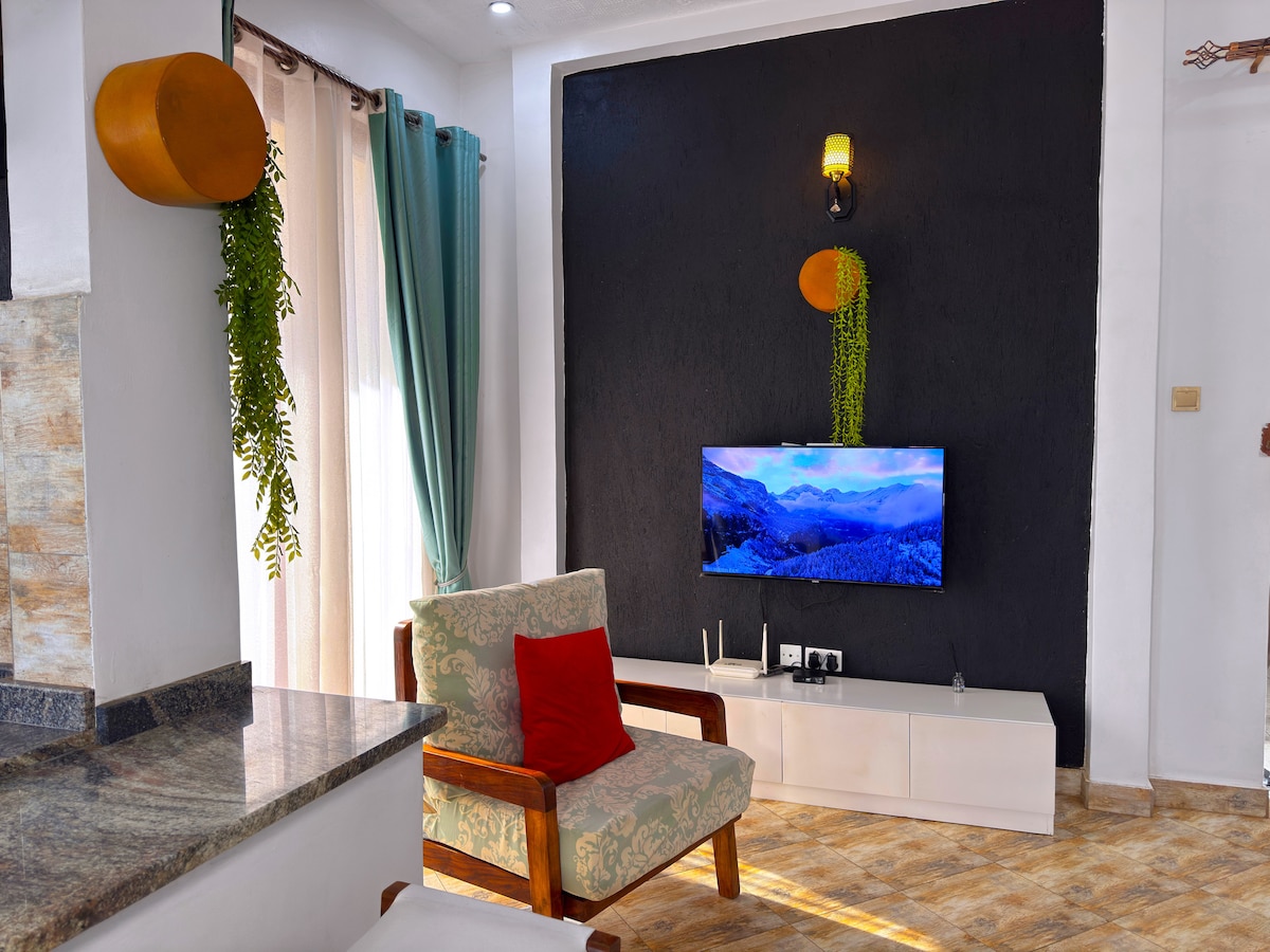 A comfortable living area features a light-colored chair with a red accent pillow, positioned near a sleek white entertainment unit. A large television is mounted on a dark accent wall, with natural light filtering through long curtains. Decorative plants add a touch of greenery.