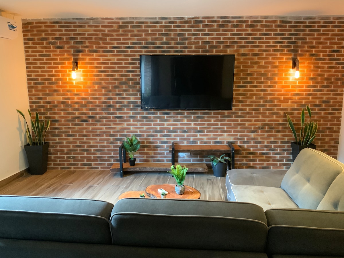 A stylish living area features a textured brick wall with two wall sconces. A large flat-screen television is mounted above a wooden shelf, while potted plants are arranged at the base. A comfortable sectional sofa and a circular wooden coffee table complete the inviting space.