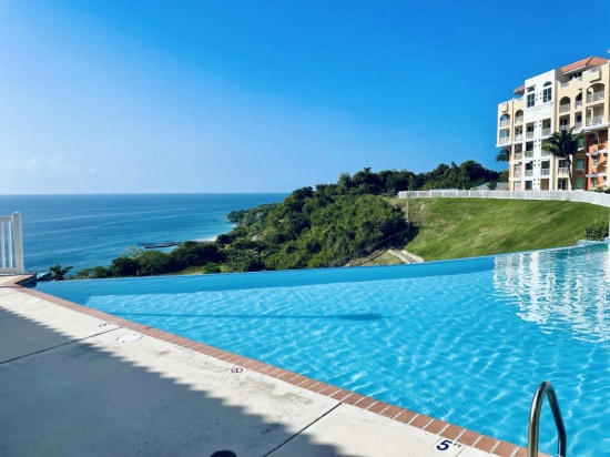 An infinity pool stretches toward the horizon, offering views of the ocean and lush greenery. The tranquil water surface reflects the clear blue sky, while the pool’s edge seamlessly merges with the landscape. A sun-soaked deck surrounds the swimming area.