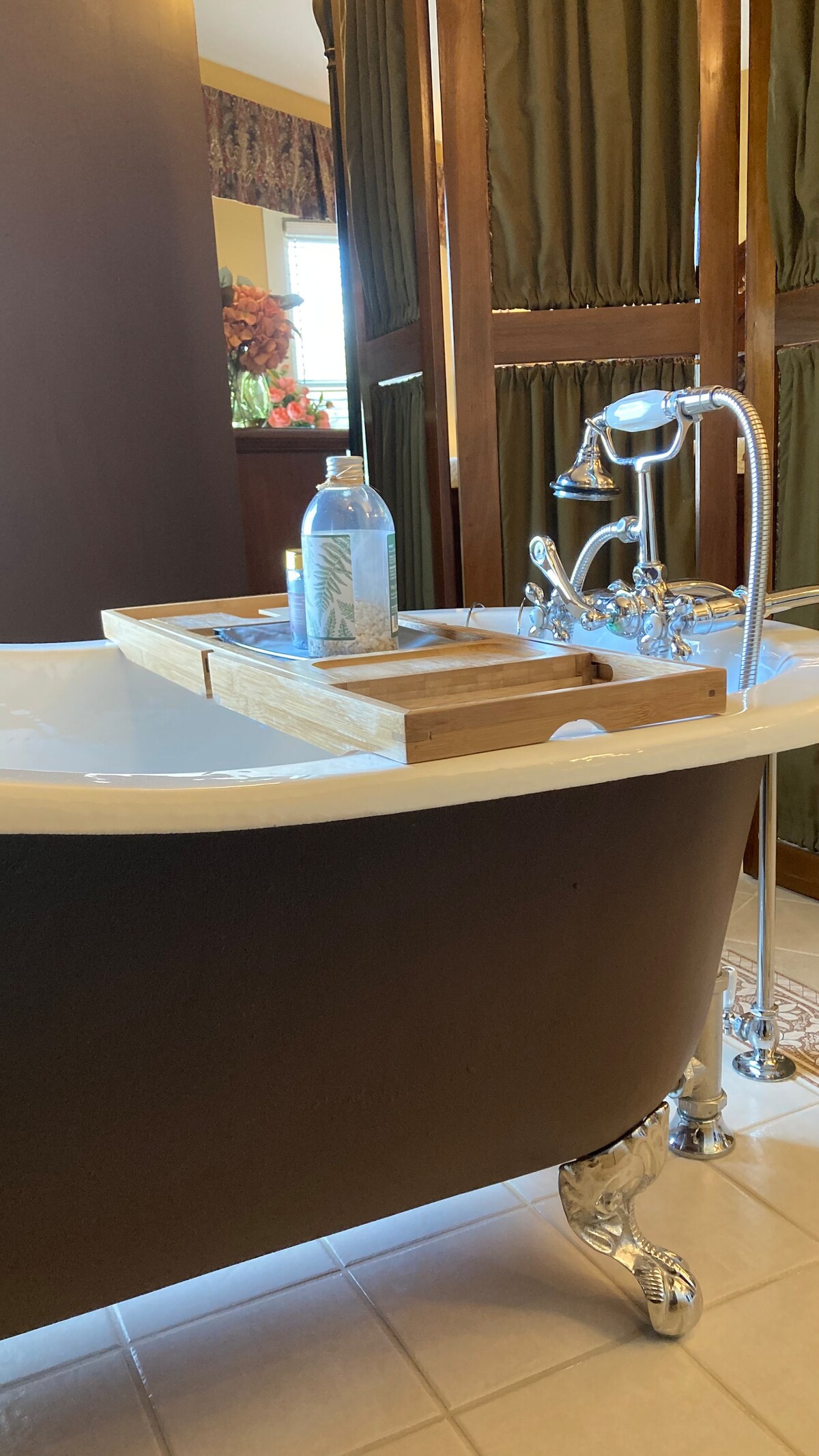 A classic freestanding bathtub is featured, complemented by a wooden caddy that holds a bottle and toiletries. The space is accentuated by rich brown paneling and curtains, with elegant chrome fixtures visible. Natural light enters the room, enhancing its relaxing ambiance.