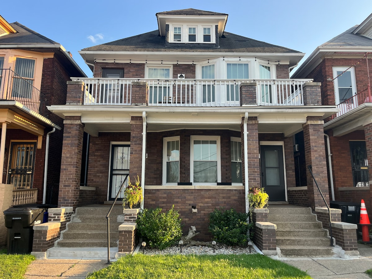 The exterior of a historic duplex features a traditional brick facade with a welcoming front porch. Steps lead to the entrance, flanked by small potted plants. Large windows provide natural light, and the surrounding greenery adds a touch of nature to the urban setting.