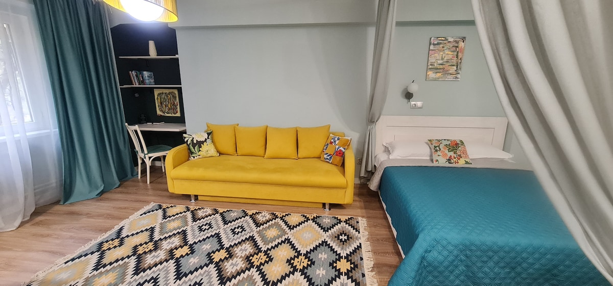 A cozy living area features a bright yellow sofa, accompanied by patterned pillows. Adjacent, a spacious bed is dressed in a teal coverlet. The floor is adorned with a geometric rug, and natural light filters through the sheer curtains of a large window.