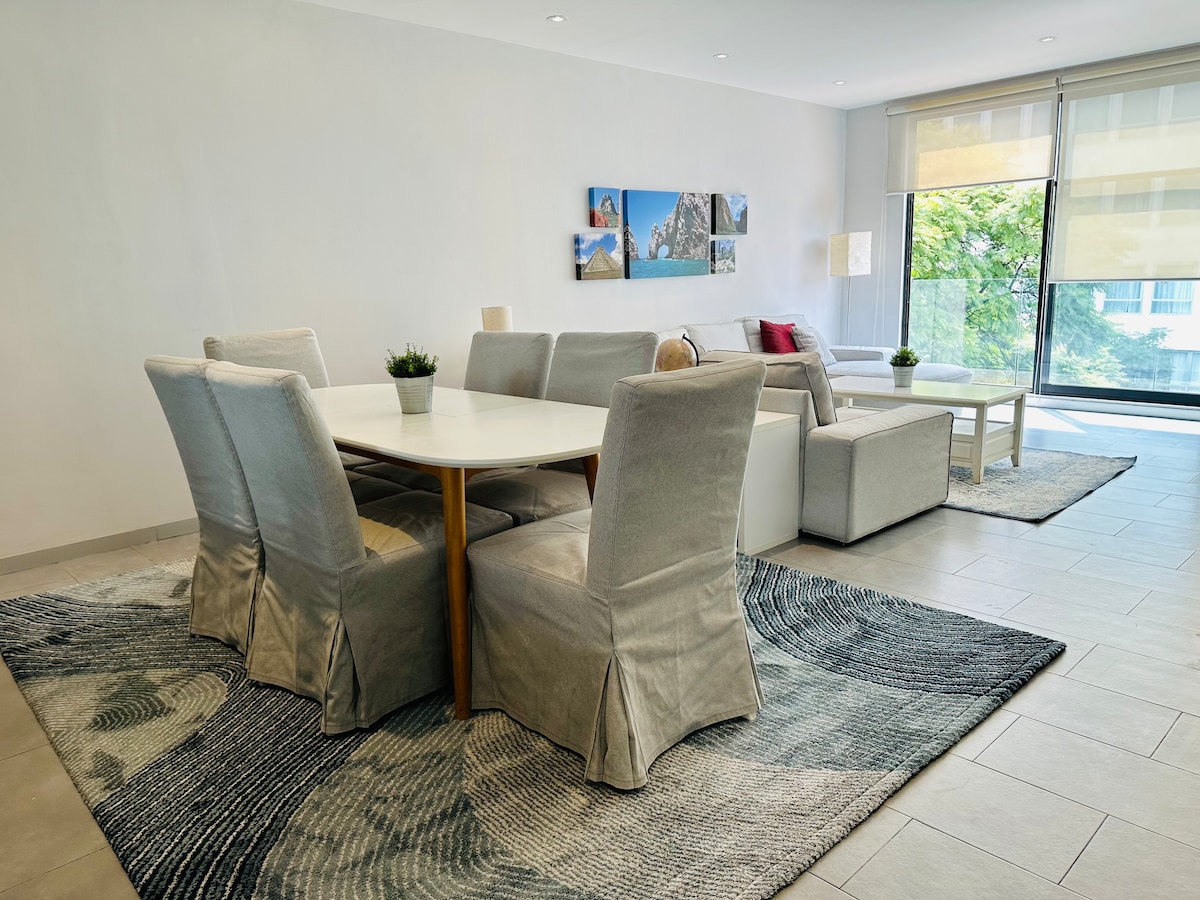 A spacious living and dining area features a light-colored dining table surrounded by six upholstered chairs. Natural light floods the space through large windows, highlighting a soft rug with a swirling design. A cozy living area with a sectional sofa and coffee table is visible, enhancing the inviting layout.