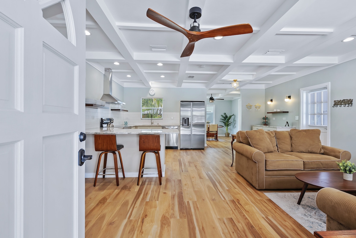 An open living area is showcased, featuring a spacious kitchen with modern appliances and a central island. Natural wood flooring flows seamlessly into a cozy seating area adorned with a comfortable sofa and accent table, creating a warm and inviting environment.