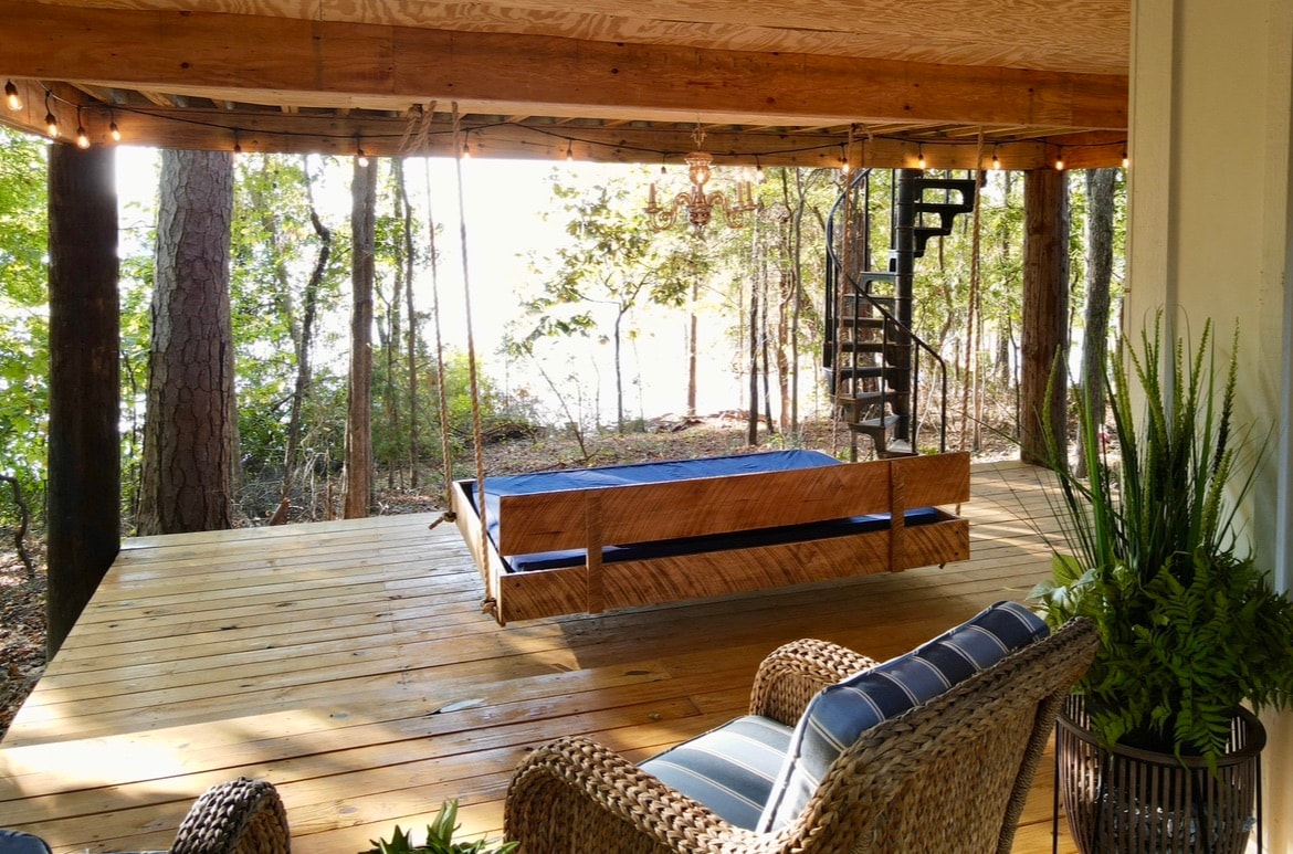 A spacious porch features a wooden swing, creating a serene space for relaxation. Comfortable seating options, including a patterned chair and a woven basket, are present. Lush greenery and trees provide a natural backdrop with views of Lake Greenwood visible in the distance.