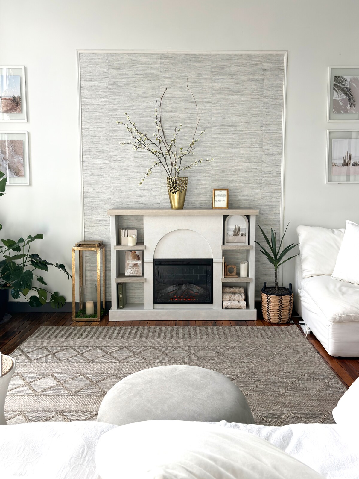A modern fireplace is positioned against a textured wall, adorned with a decorative vase and branches. Shelves display curated items, complemented by a wicker basket and plants. A large area rug anchors the space, with light-colored furniture arranged for comfort.