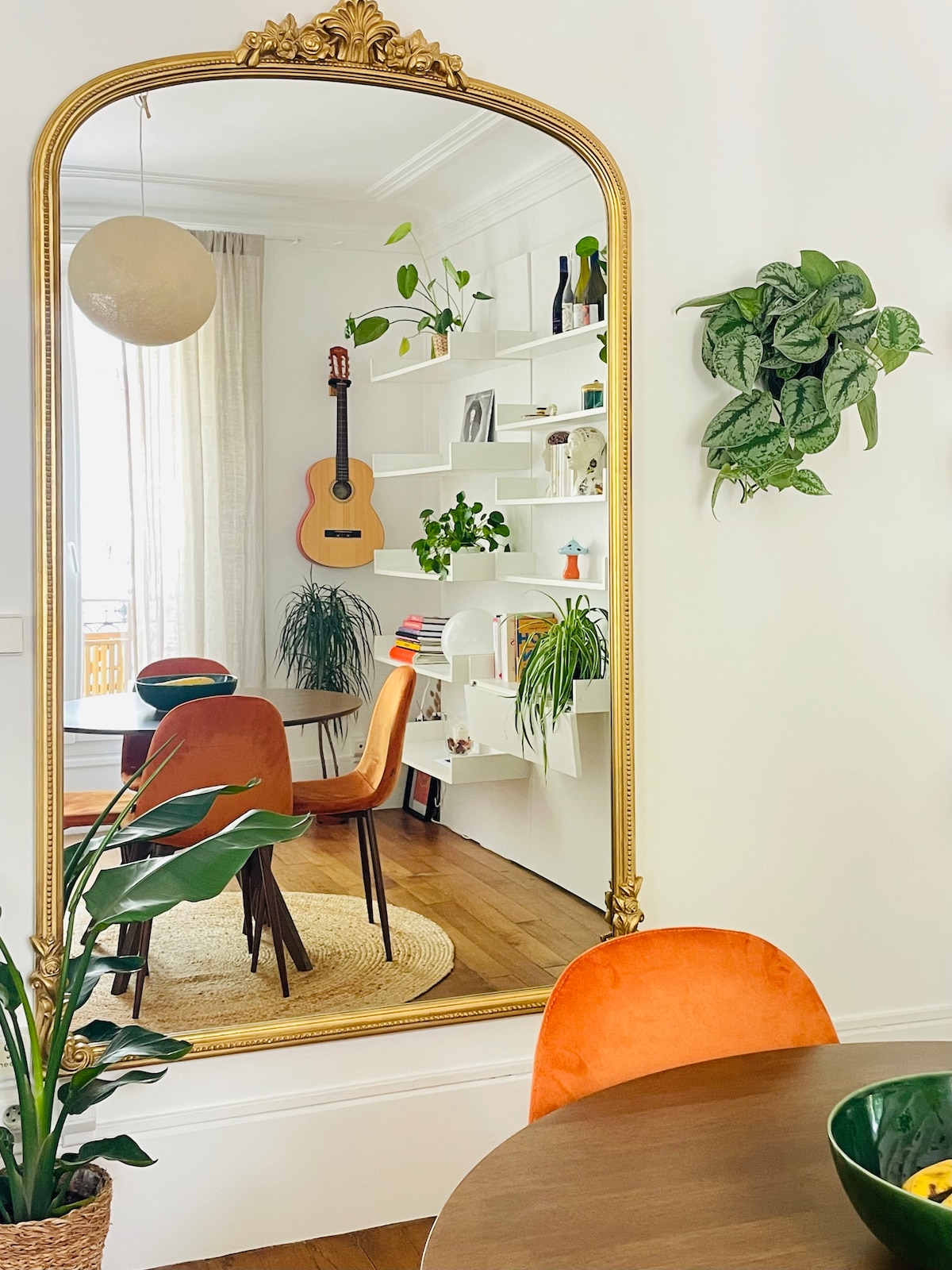 A large, ornate mirror reflects a cozy dining area featuring colorful chairs and a wooden table. Plants adorn the wall shelves and nearby surfaces, contributing to a vibrant atmosphere. Soft natural light filters through a nearby window, enhancing the inviting space.