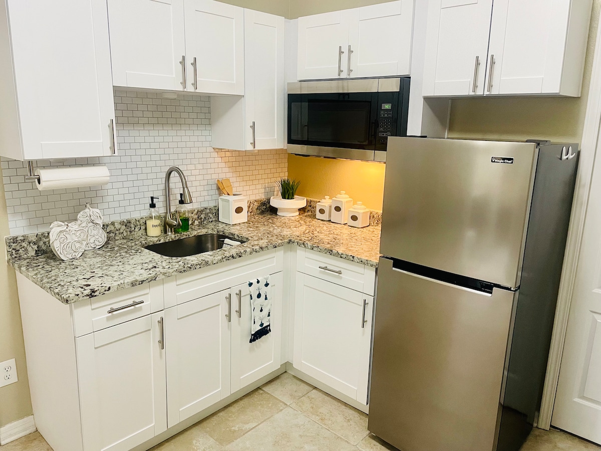 A modern kitchen is equipped with white cabinetry and a polished granite countertop. Stainless steel appliances, including a microwave and refrigerator, complement the design. A small sink is situated beneath a window, and decorative elements are placed on the countertop.