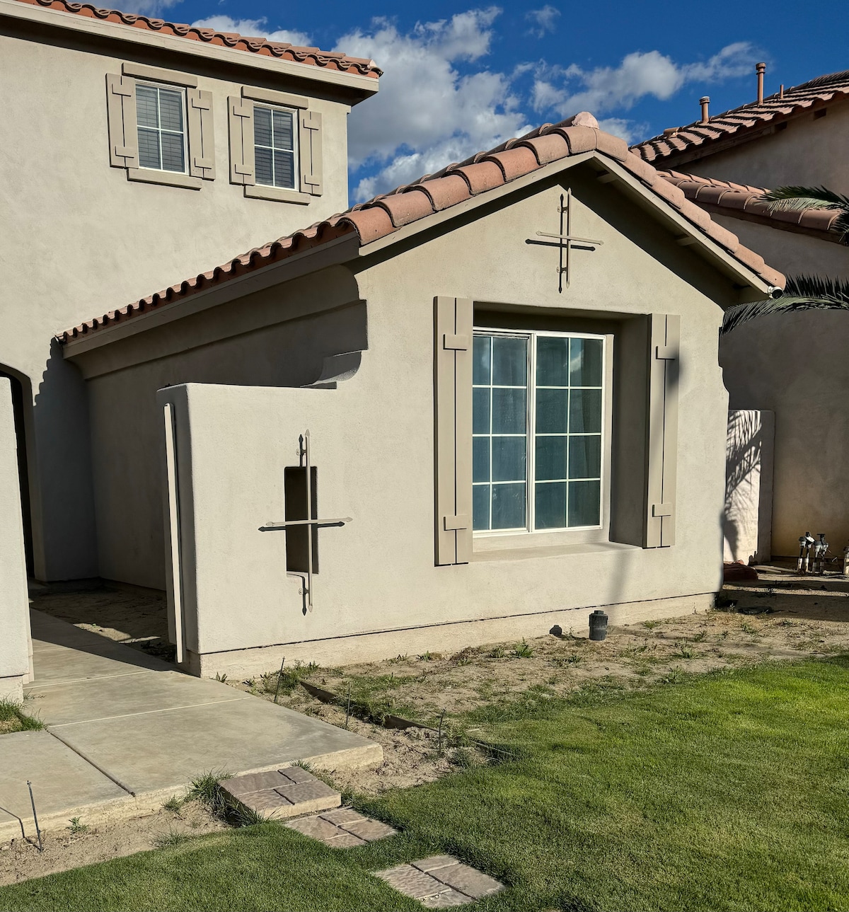 The exterior of the casita is showcased, featuring a light-colored stucco facade and a distinctive, large window framed by decorative shutters. The landscape includes trimmed grass and a pathway leading to the entrance, under a partly cloudy sky.