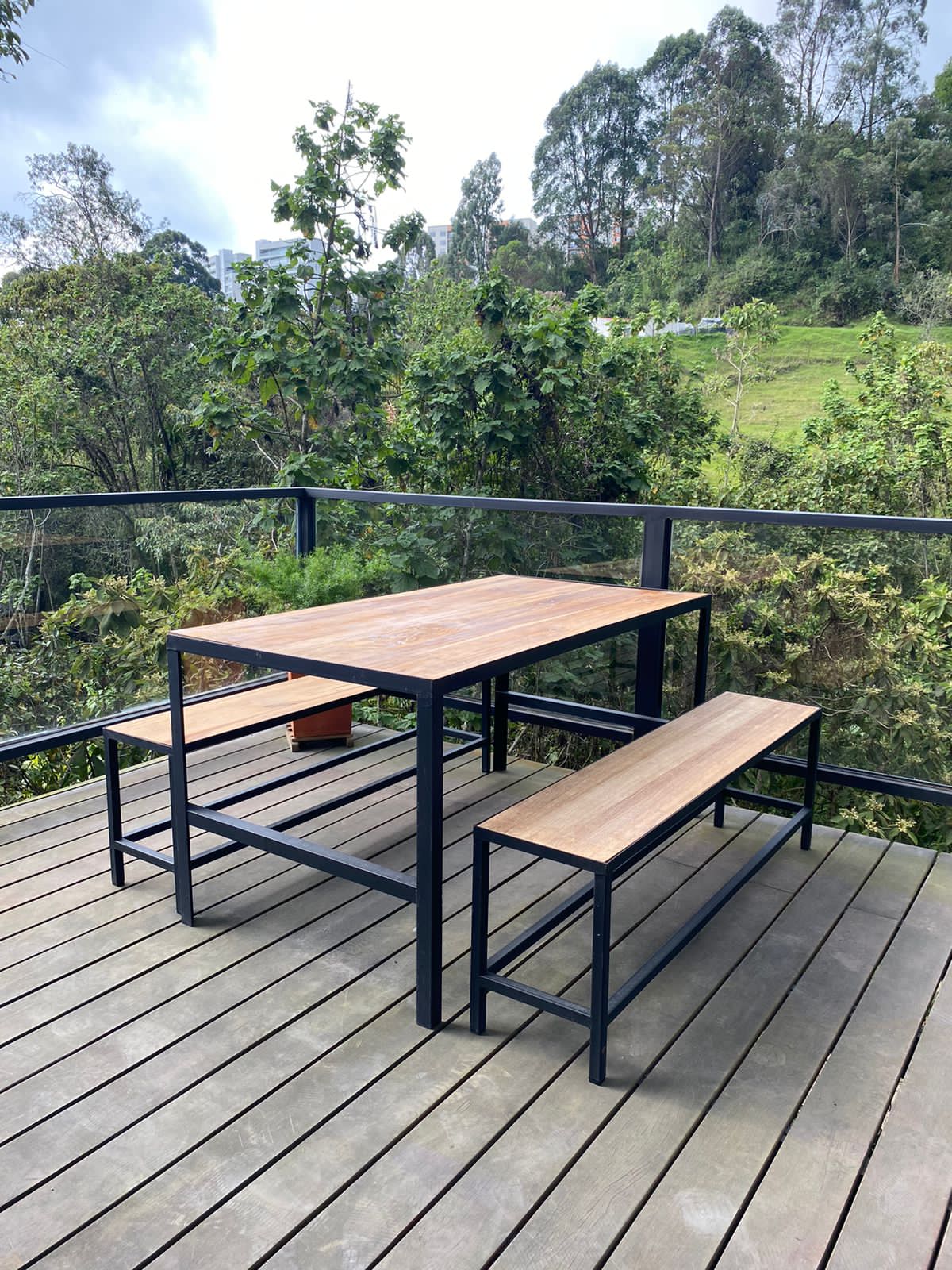 An outdoor terrace features a wooden table accompanied by two benches, all resting on a wooden deck. Lush greenery and trees are visible in the background, contributing to a serene natural setting.