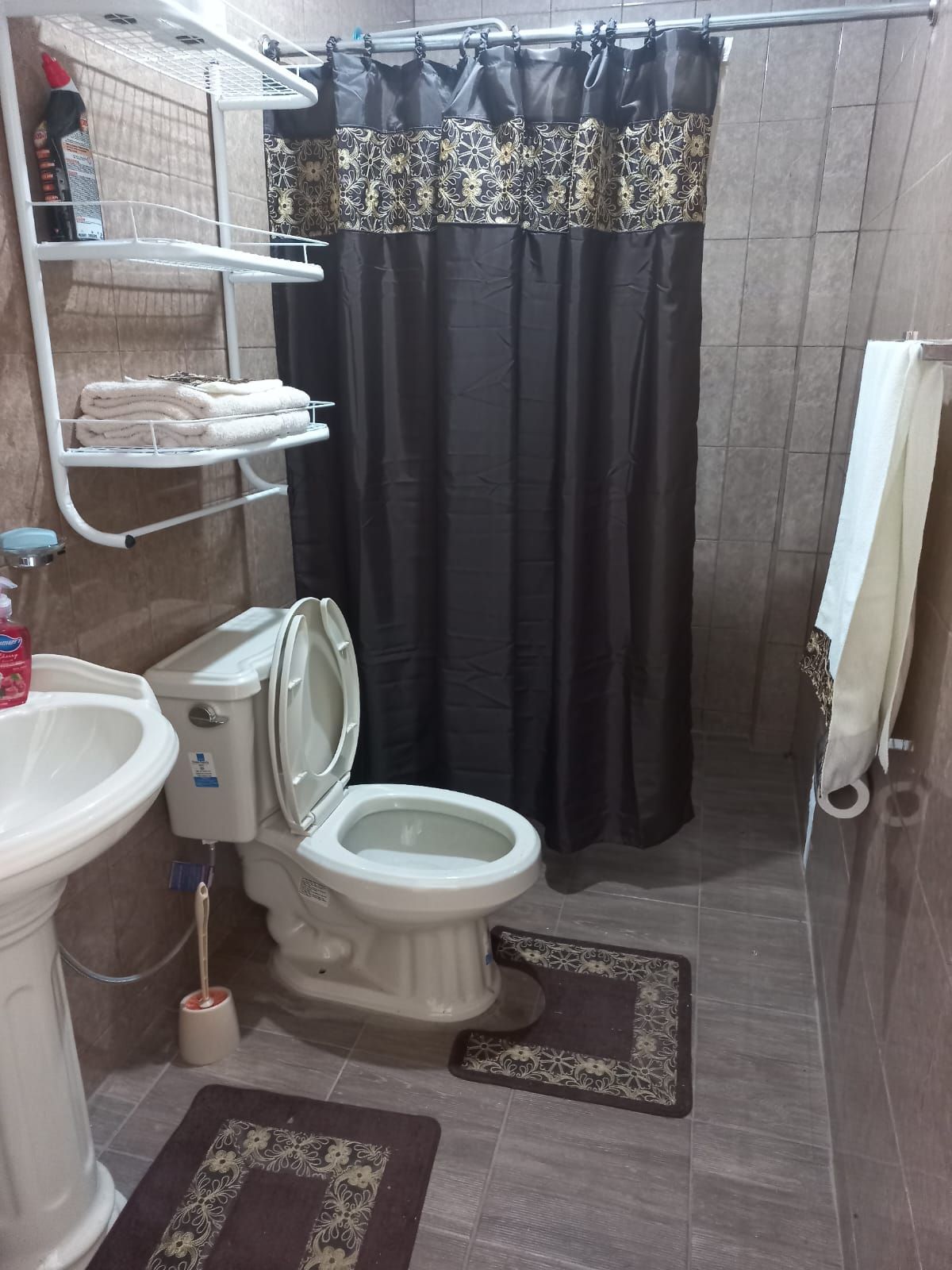 A compact bathroom is highlighted by neutral tile walls and a matching floor. A white sink and toilet are positioned thoughtfully. A shower curtain adds a touch of elegance, while neatly arranged towels are displayed on a hanging rack. A toilet mat complements the overall decor.