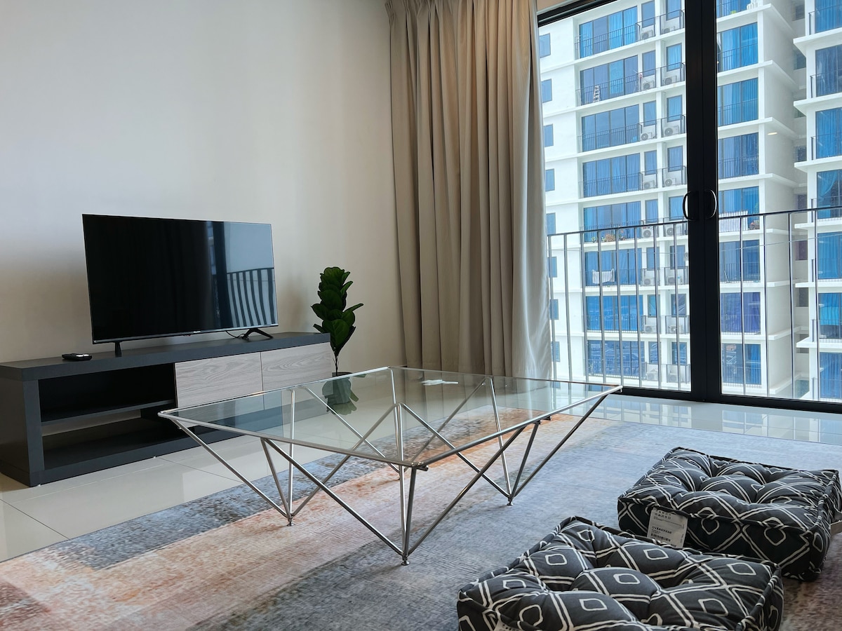 A modern living space is highlighted by a geometric glass coffee table, complemented by patterned cushions. A television is positioned on a sleek stand, with large windows offering natural light and views of the surrounding architecture.