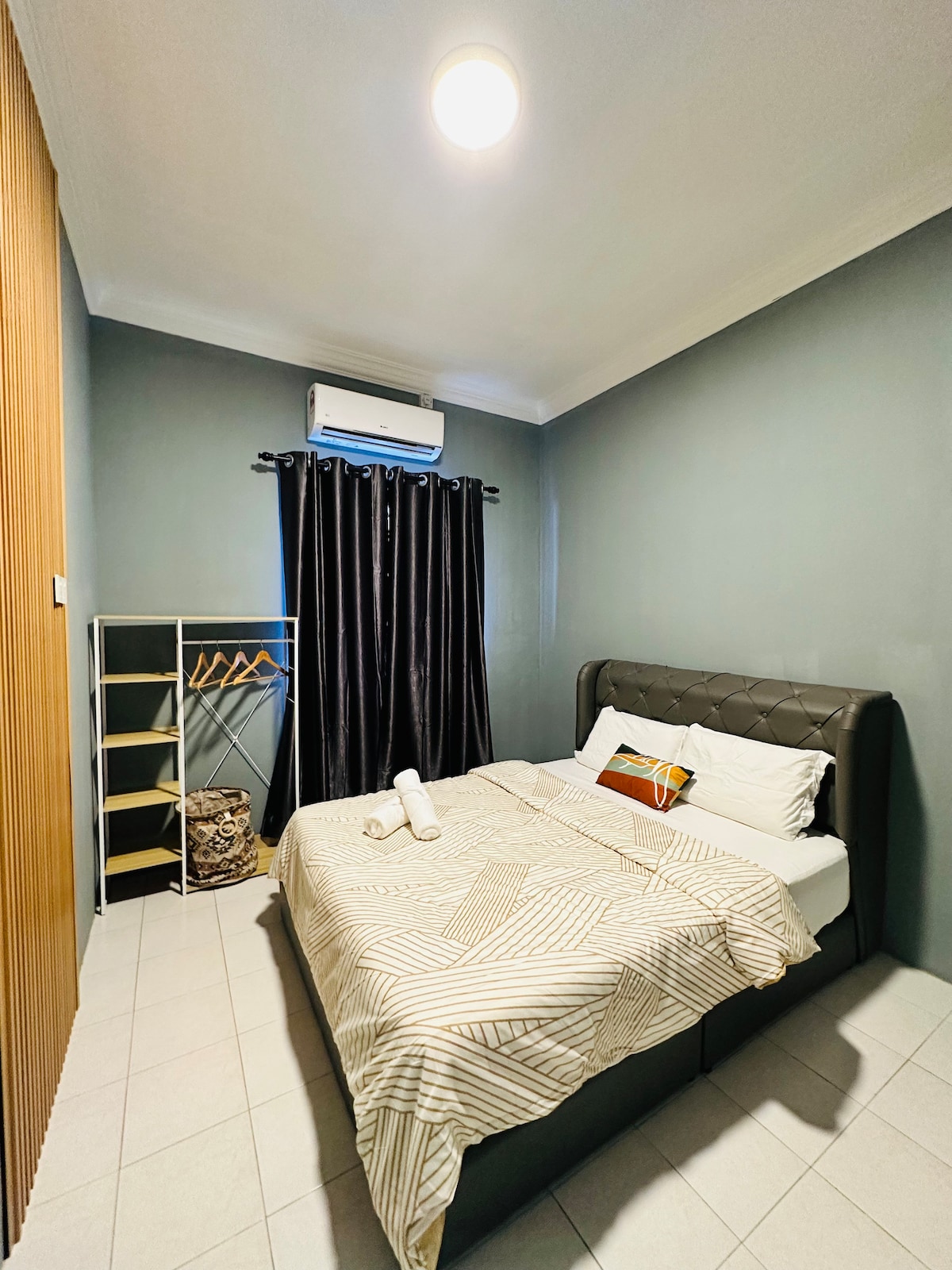 A cozy bedroom features a dark accent wall and a comfortable bed dressed in a geometric-patterned comforter. A small wardrobe with open shelving is present, along with a curtain that covers a window. Air conditioning is mounted on the wall for climate control.