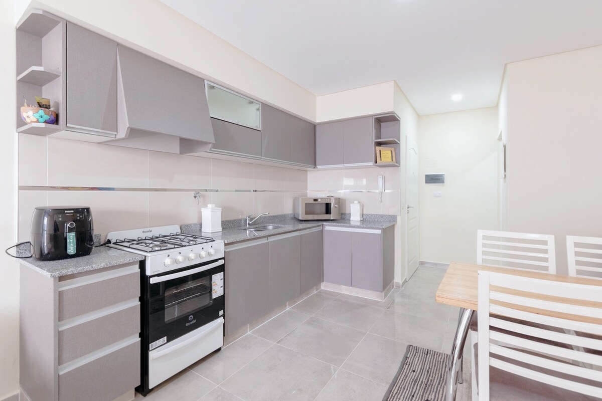 A modern kitchen is showcased with sleek gray cabinetry and light countertops. A gas stove and microwave are visible, along with a small countertop appliance. A wooden dining table with white chairs is present, creating a functional space for meals.