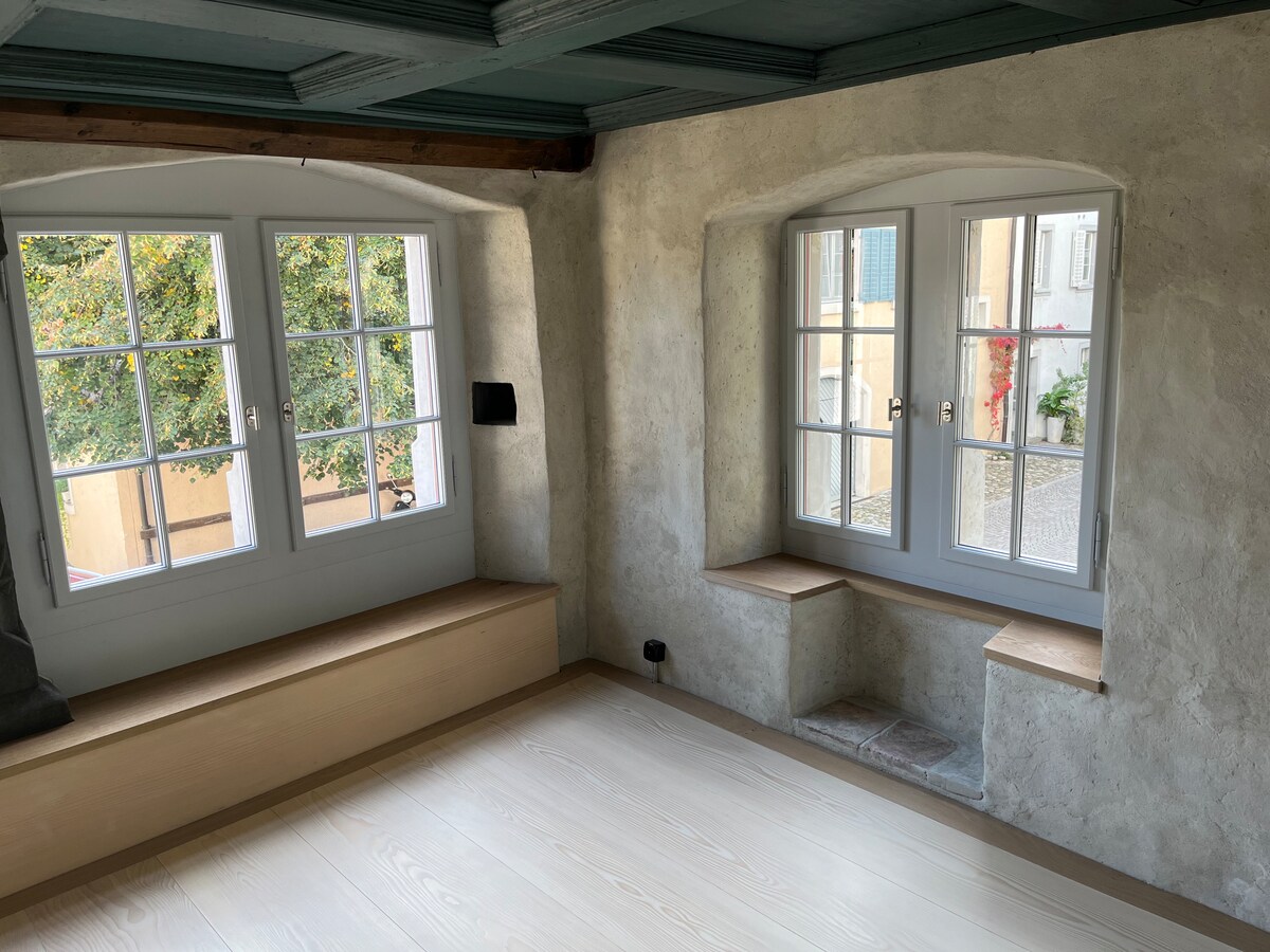 Two large windows are framed in white, allowing natural light to fill the spacious room. Each window features a wooden sill, and the light-colored wooden floor complements the soft, neutral tones of the walls, which exhibit a textured finish.