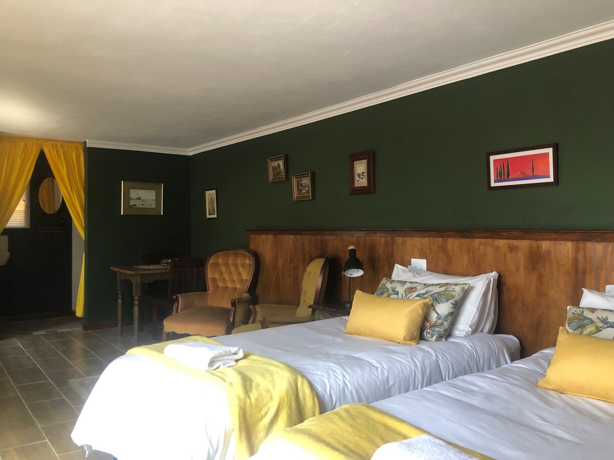 A spacious room features two neatly arranged beds adorned with yellow accents. The green walls create a calm environment, complemented by wooden paneling. A comfortable armchair sits in one corner, and tasteful artwork adorns the walls, enhancing the inviting decor.