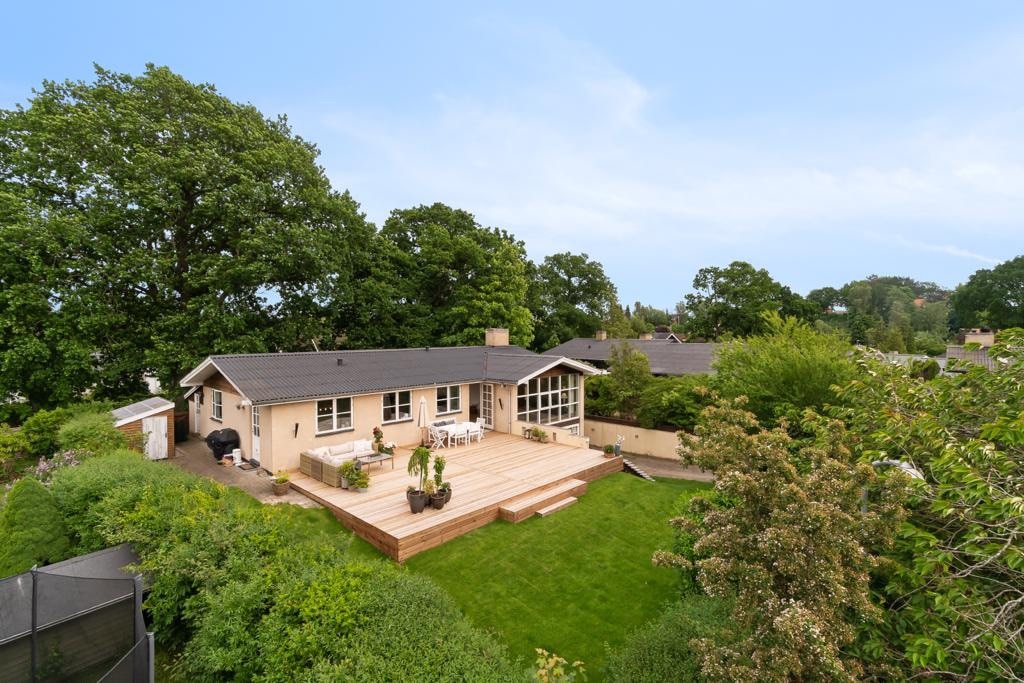 A spacious backyard is framed by lush greenery, featuring a wooden deck that extends from the house. The exterior of the home is light-colored with large windows, allowing natural light to brighten the interior. A gas grill is positioned near the deck.