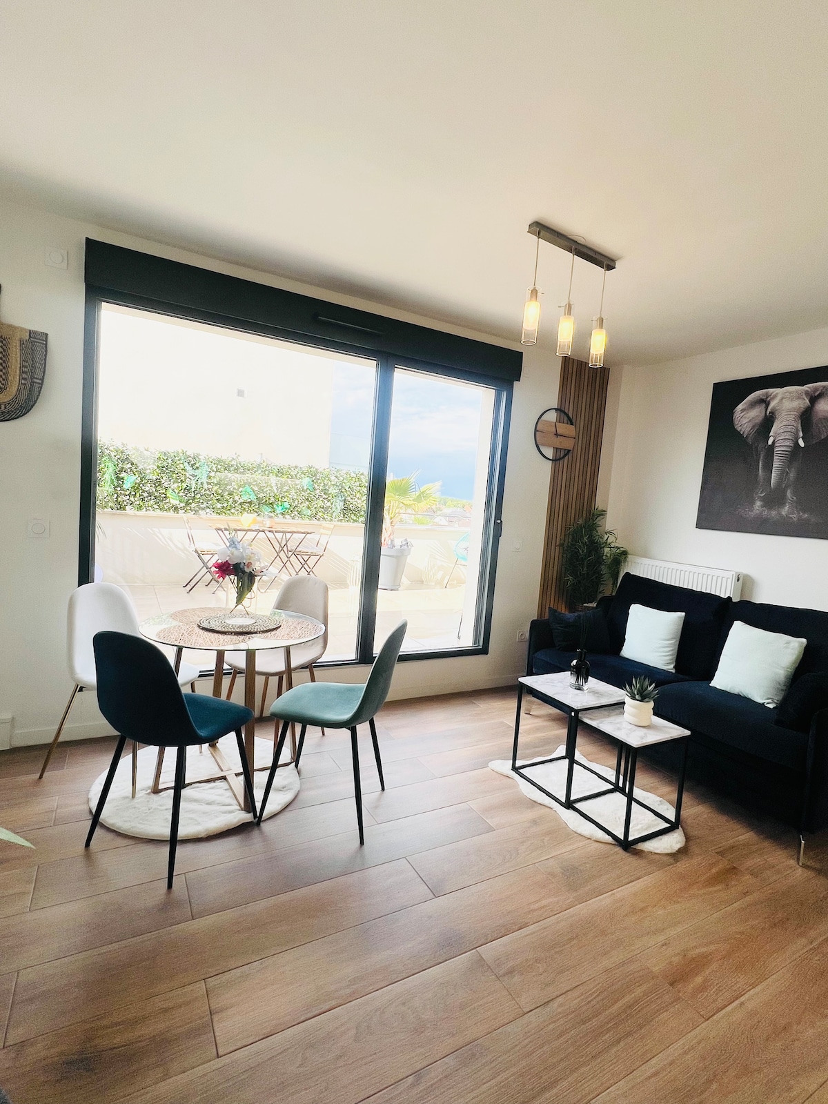 A modern living area is visible, featuring a sleek black sofa and a small round table with chairs. Natural light floods the space from large glass doors leading to an outdoor terrace. Decorative elements include a floral arrangement and wall art featuring an elephant.