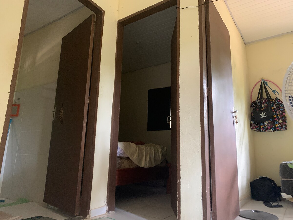 Two wooden doors are shown, leading to separate rooms. A glimpse of a spacious bed with light bedding is visible through one door. The hallway area features neutral-colored walls and tile flooring, contributing to a simple and functional living space.