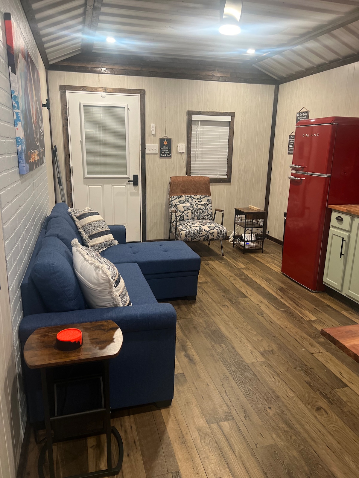 A cozy living area features a blue sectional sofa and a red refrigerator, complemented by wooden flooring. A wooden table with a small orange object adds functionality. Nearby, a patterned chair enhances comfort. Natural light enters through a door and window, creating an inviting space.