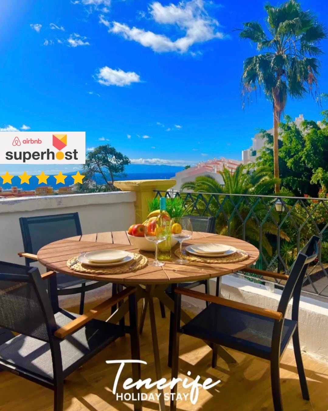 A round table with two plates and a bowl of fruit is set on the terrace, surrounded by four chairs. Lush greenery and palm trees are visible in the background, along with a view of the Atlantic Ocean under a clear blue sky.