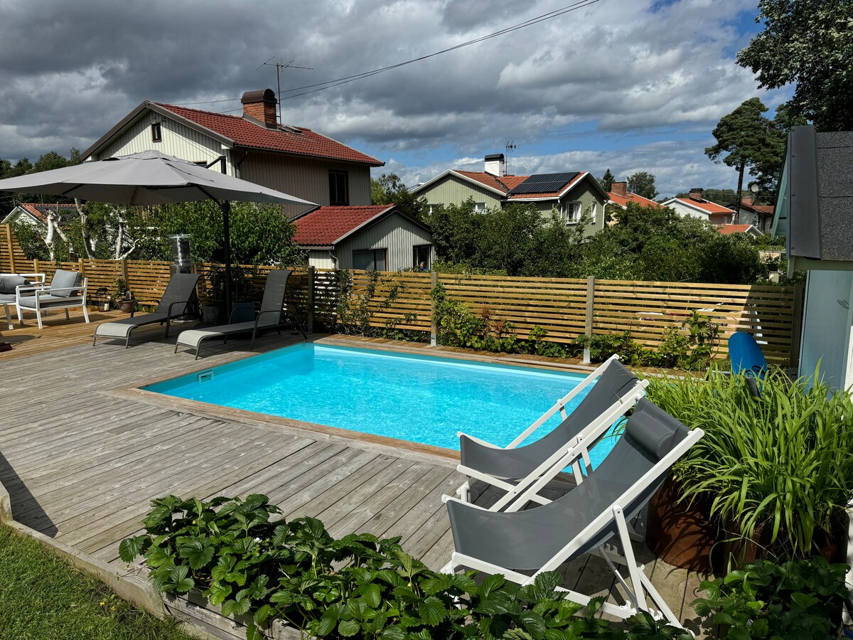 A heated pool is framed by a wooden deck, featuring two lounge chairs and an umbrella for shade. Lush greenery and a wooden fence provide privacy, while the surrounding area displays charming houses under a partly cloudy sky.