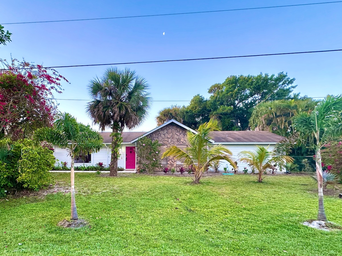A charming single-family home is surrounded by lush greenery and vibrant landscaping. The front entrance features a striking pink door and stone accents. Palm trees and flowering plants provide a tropical feel, while the well-maintained lawn enhances the inviting exterior.