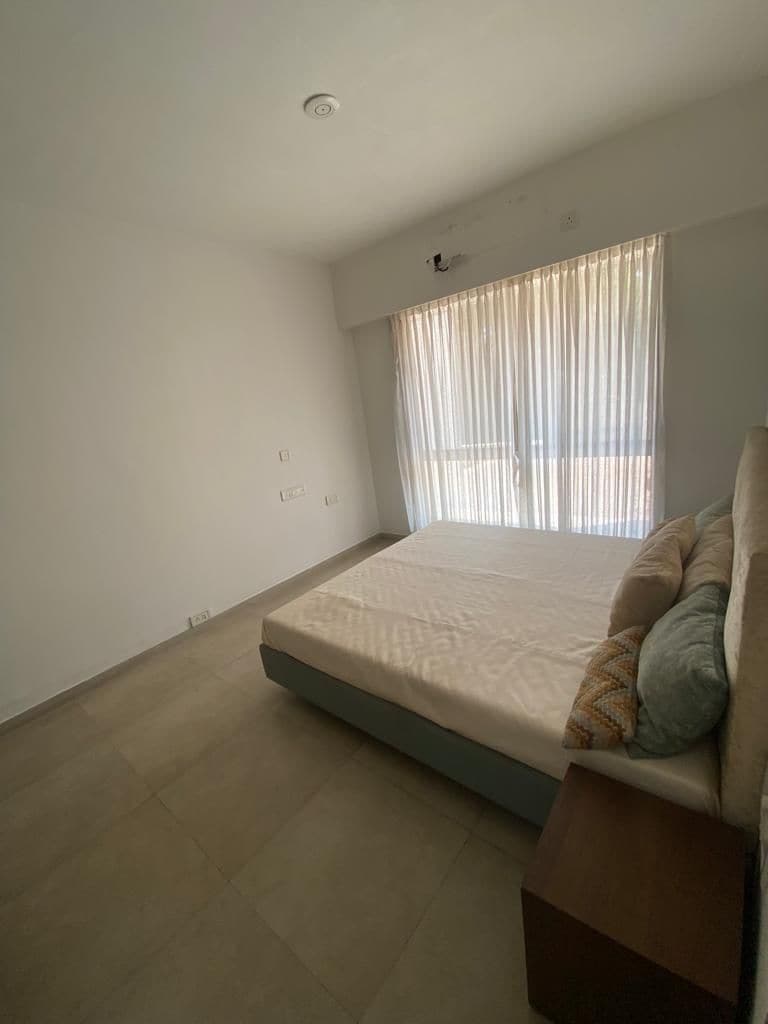 A bright bedroom features a spacious bed with neutral bedding. Natural light filters through sheer curtains covering a wide window, illuminating the pale tiled floor. A wooden nightstand is positioned beside the bed, enhancing the room's minimalist design.