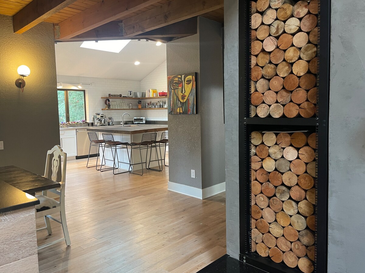 The image showcases a view of a spacious kitchen area featuring modern appliances and a large island with ample seating. A decorative wall-mounted storage unit displays neatly stacked firewood beside an artwork piece, adding a touch of character to the open layout.