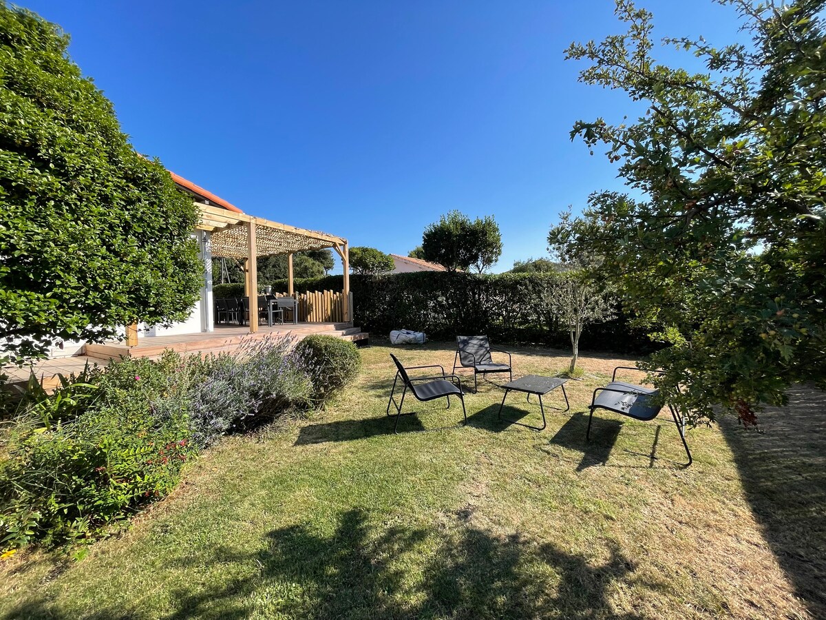 A spacious garden is surrounded by neatly trimmed hedges, offering an area with several lounge chairs. A shaded patio can be seen in the background, which provides a relaxing spot for outdoor dining. The serene blue sky creates a calm atmosphere over the landscape.