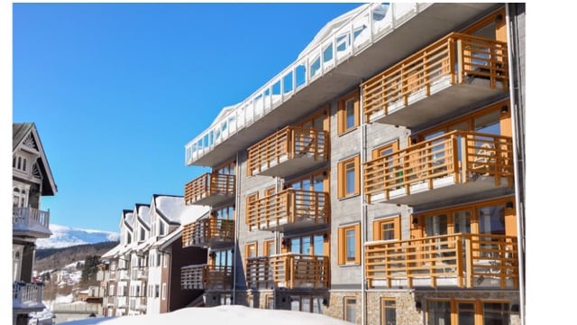 The image shows a multi-level building with a modern architectural design, featuring wooden balconies on each floor. The facade is a combination of gray and orange hues, set against a clear blue sky. Snow can be seen on the ground, highlighting the winter setting.