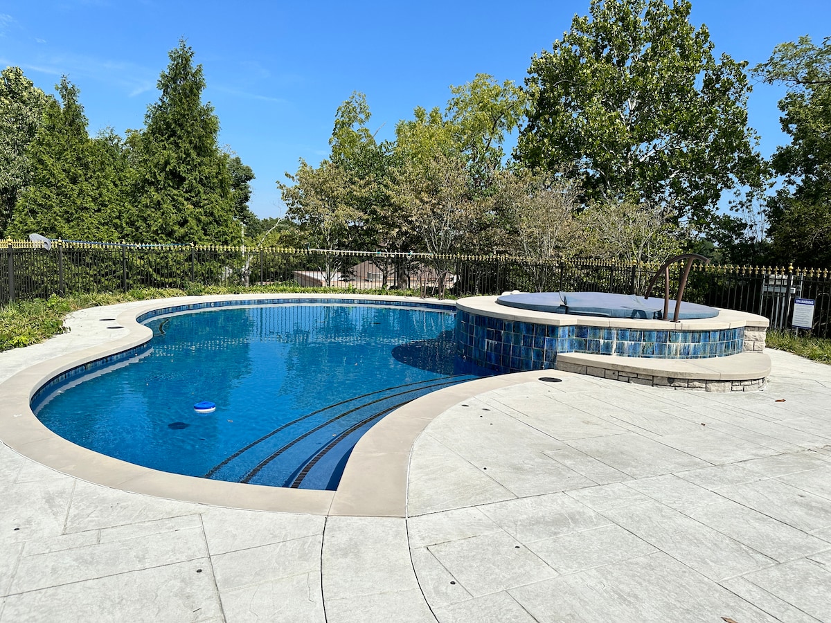 A curved pool with crystal-clear water is surrounded by a manicured landscape. The area features a hot tub adjacent to the pool, and a black wrought iron fence encloses the space. Trees provide partial shade, while a blue sky enhances the tranquil setting.